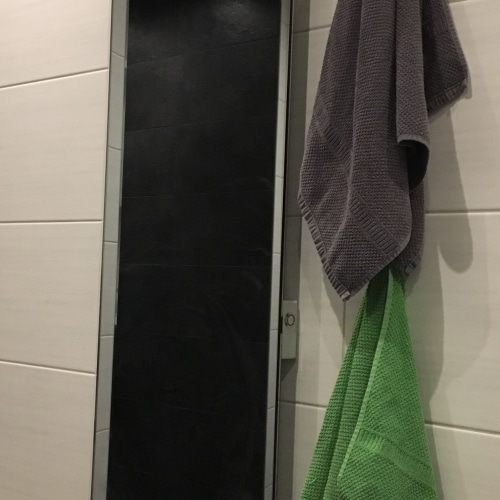 Ein schwarzer Handtuchtrockner aus Glas in einem Badezimmer mit weißen Fliesen, wo daneben ein graues und ein grünes Handtuch hängen.